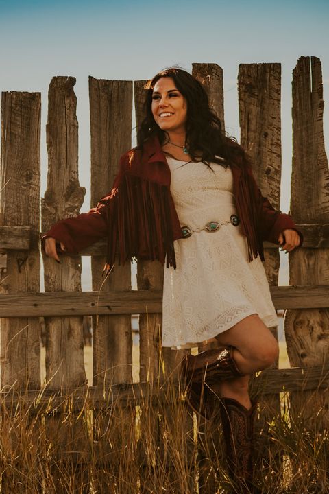 Ashley Sara wearing a fringe jacket and cowboy boots, leaning on a wooden fence at sunset
